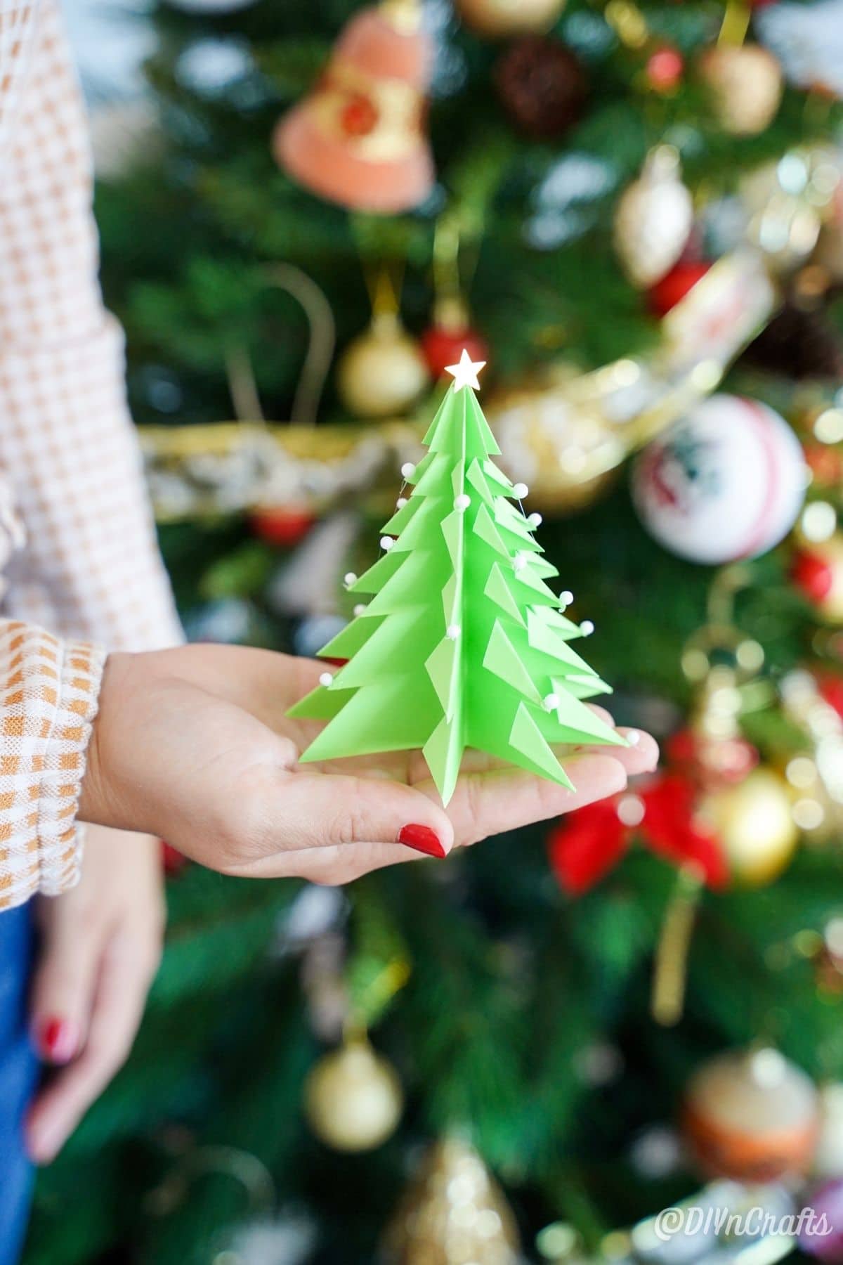 женщина держит зеленую бумажную елку перед украшенной рождественской елкой