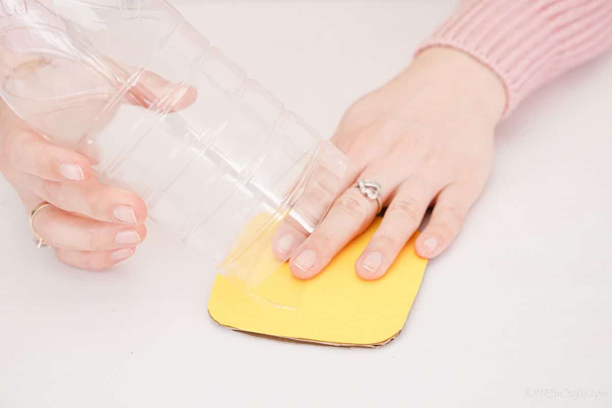 рука держит желтый картонный квадрат под пластиковой бутылкой